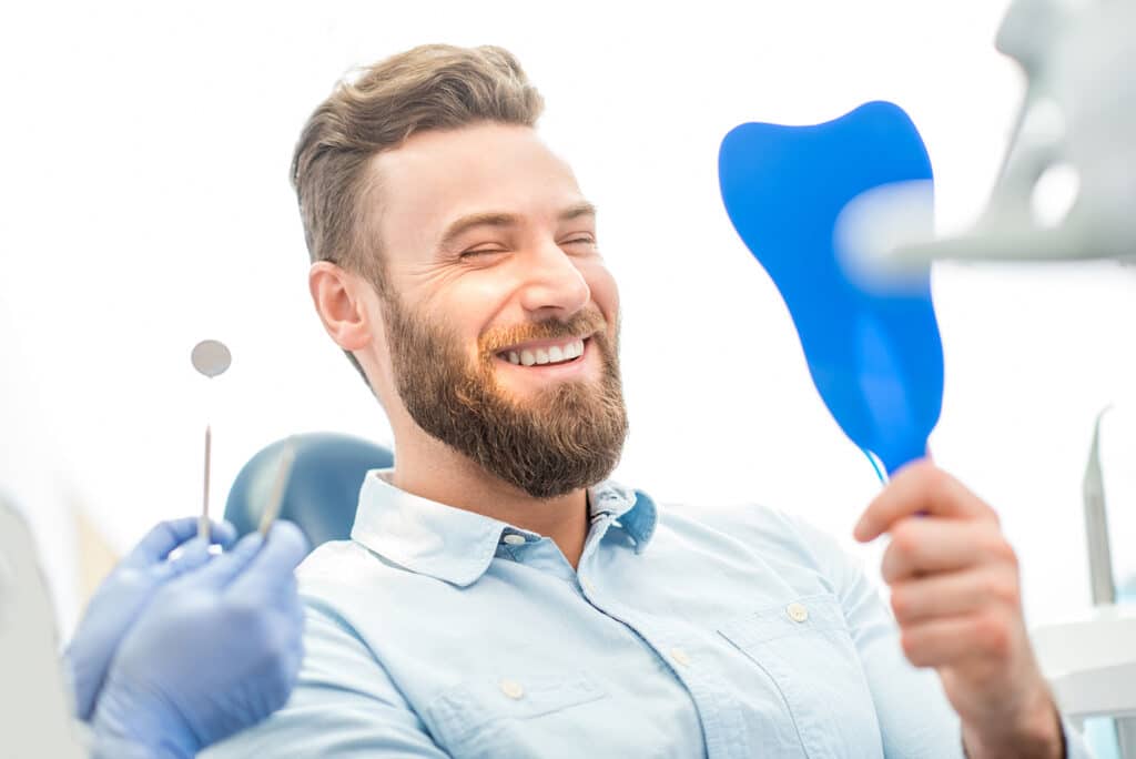 A man smiling while holding a mirror after dental treatment